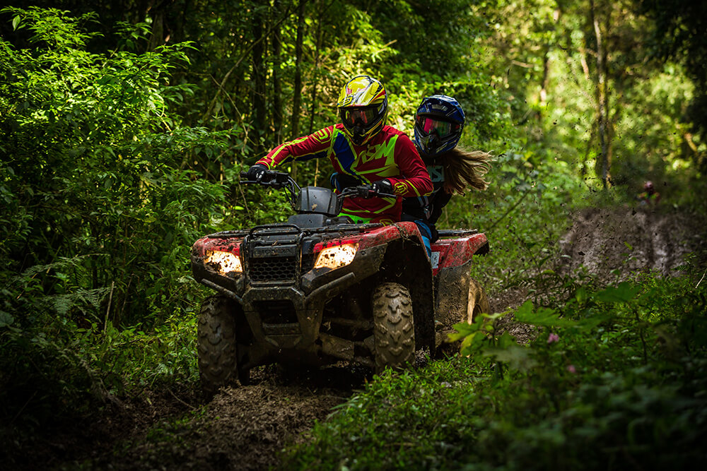 Two people ride on a red ATV in the forest wearing full protective gear.