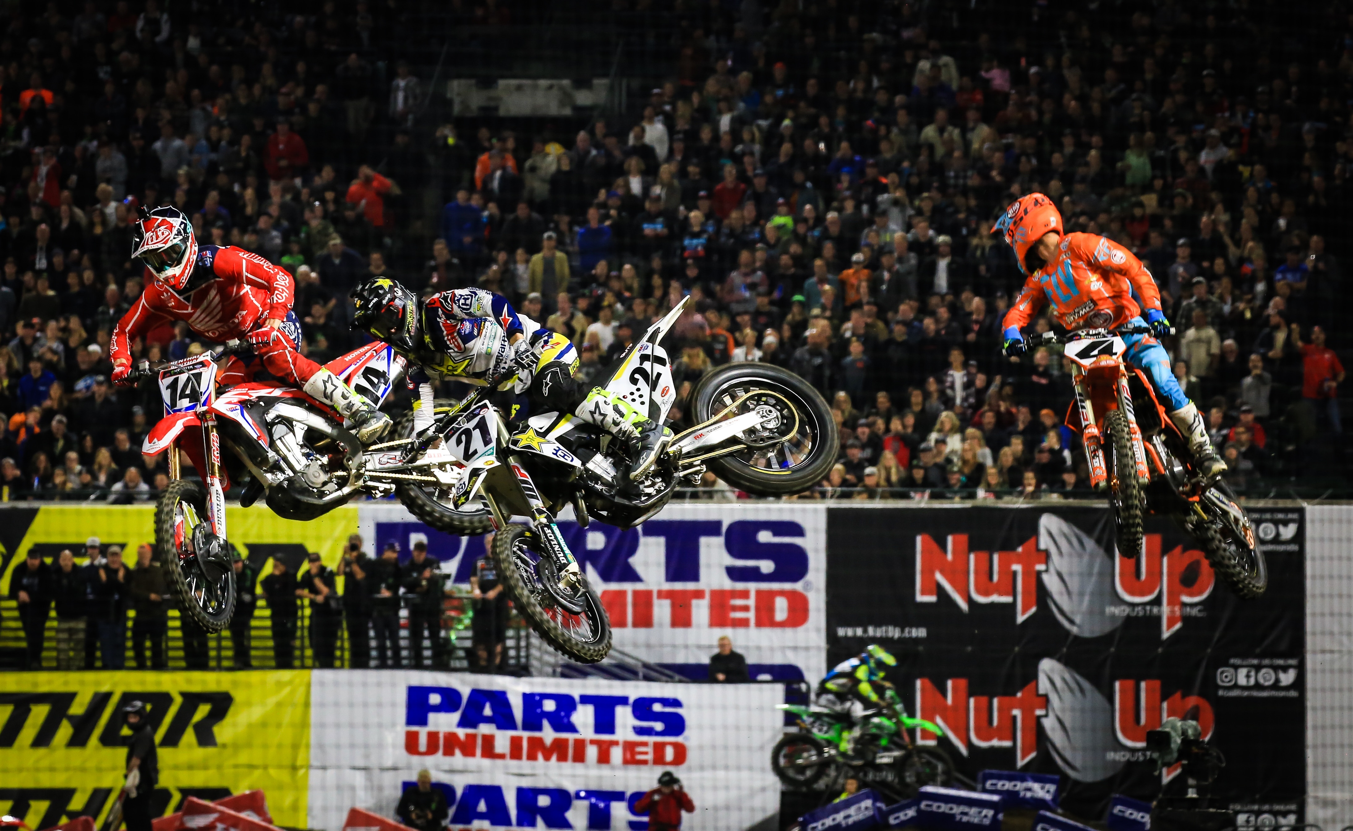 Three dirt bike riders fly through the air as they race around a track with packed stands.