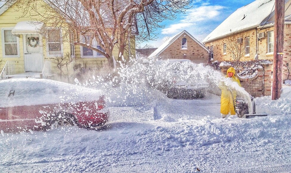 man wearing yellow snow suit clears snow from his driveway with a snowblower